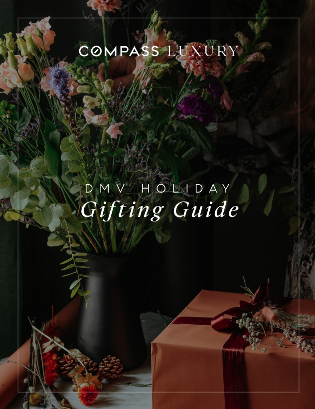 A floral arrangement in a black vase next to a wrapped gift box with a maroon ribbon, on a table with pinecones and decorative items. Overlaid text reads 'Compass Luxury DMV Holiday Gifting Guide.'