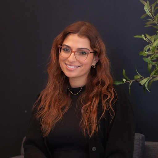 A woman with long wavy red hair wearing glasses and a black top, sitting in front of a dark background and some green foliage.
