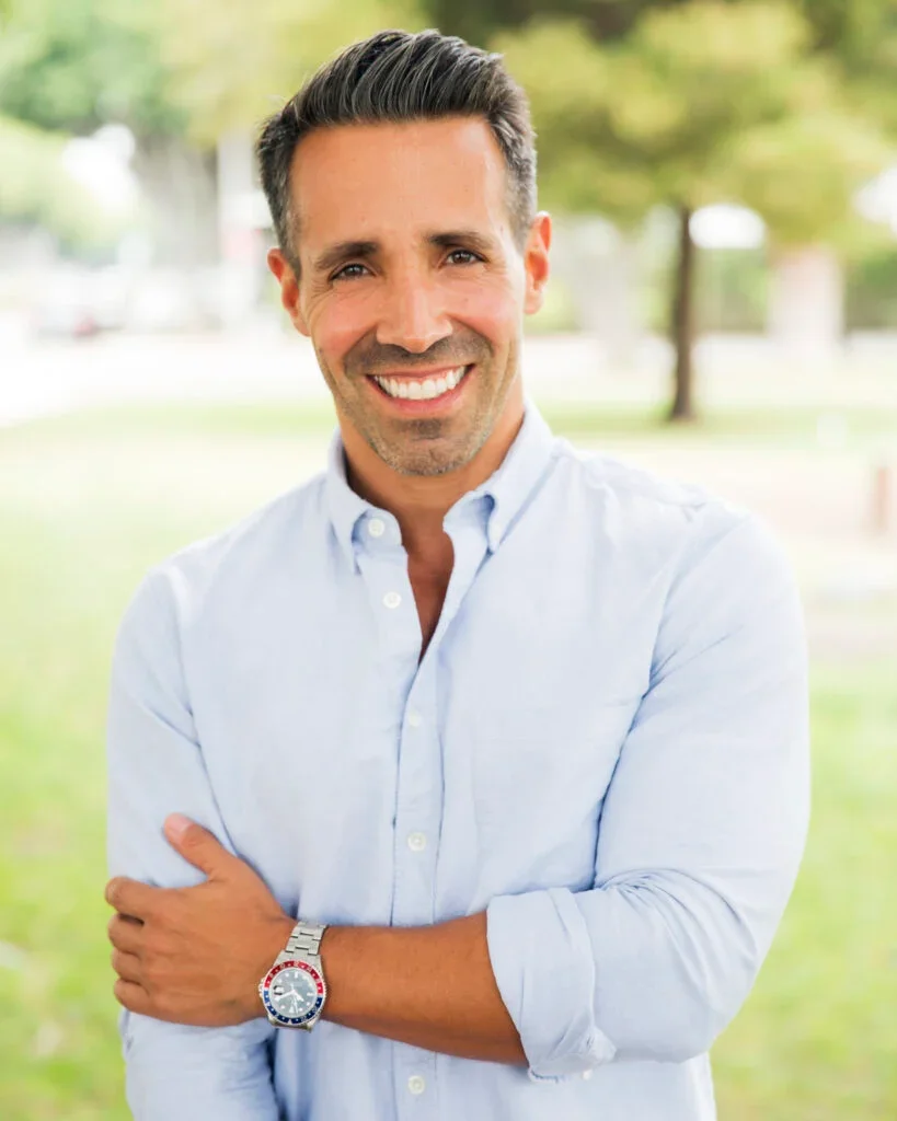 Smiling man with short dark hair, wearing a light blue button-up shirt, standing outdoors with trees and a park in the background.
