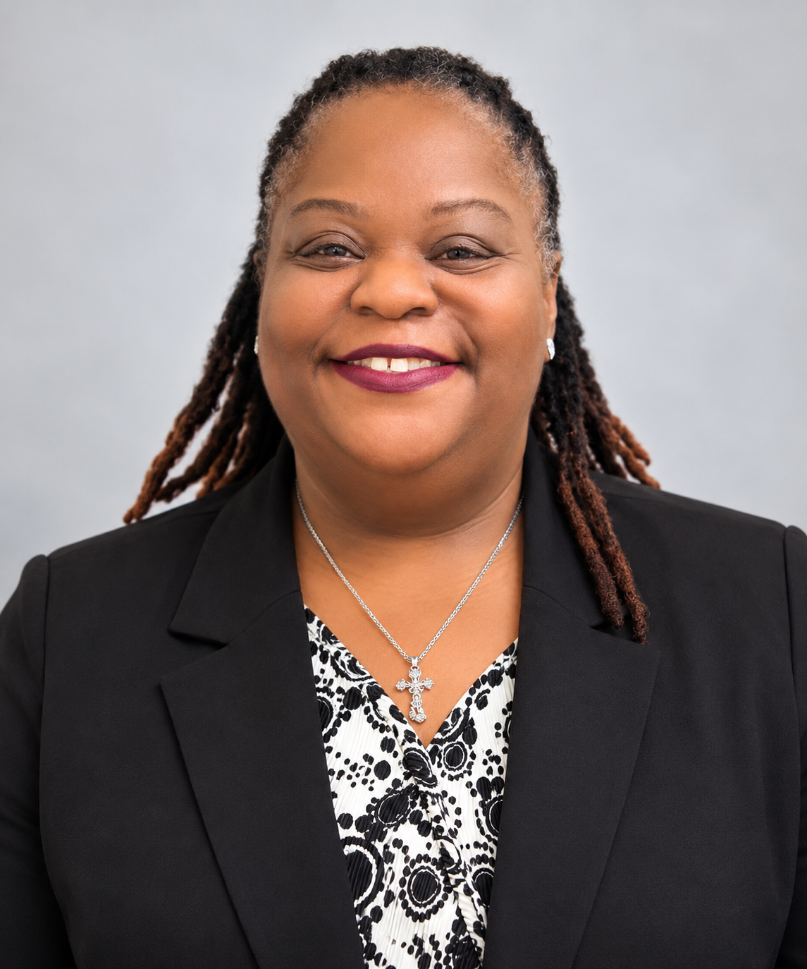 A smiling woman with dark skin and dreadlocks wearing a patterned blouse, a vest, and a cross necklace, standing against a plain beige wall.