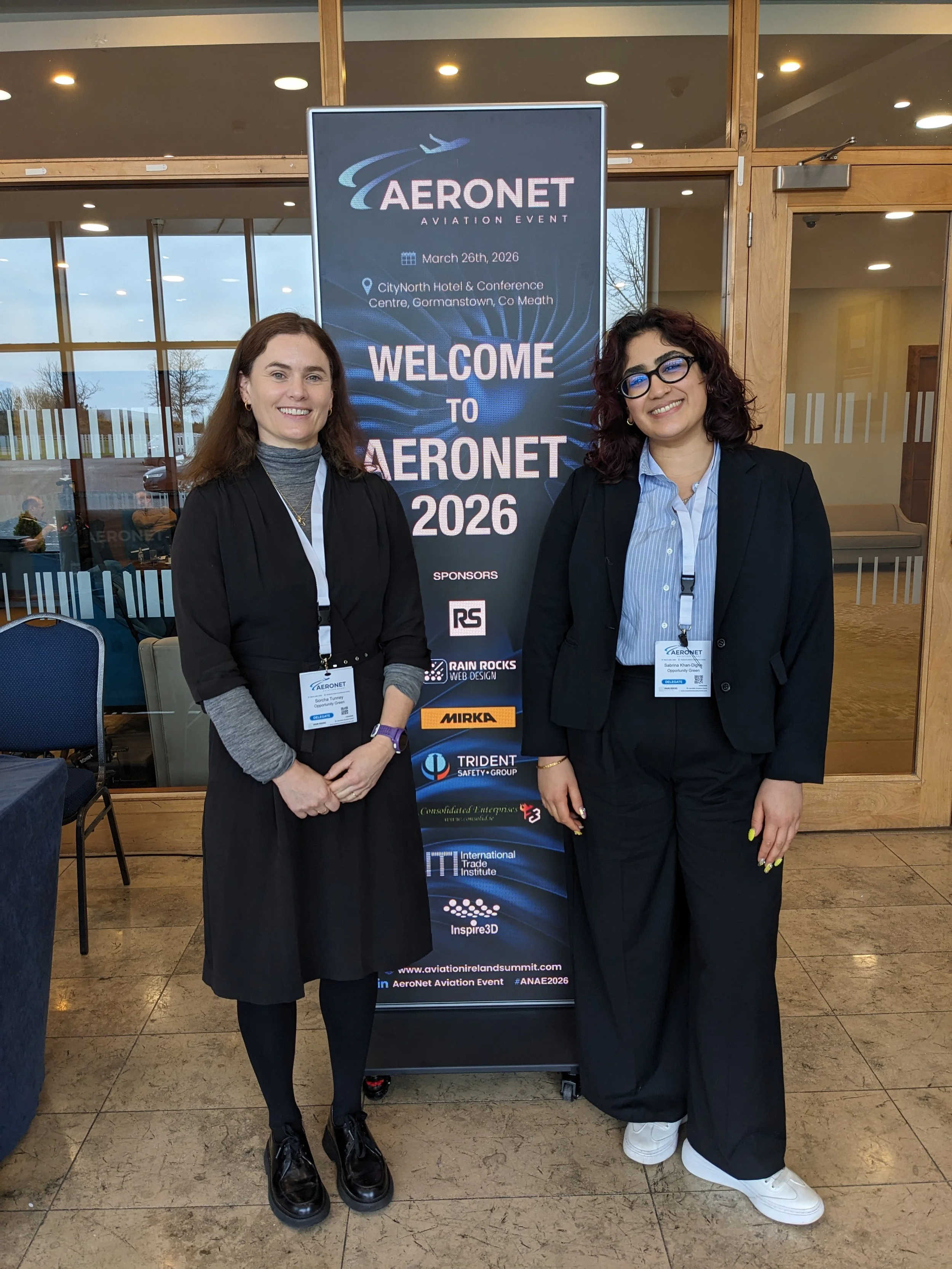 Two women smiling with a banner that says "Aeronet Aviation Event" between them. They are dressed smart and have lanyards on.
