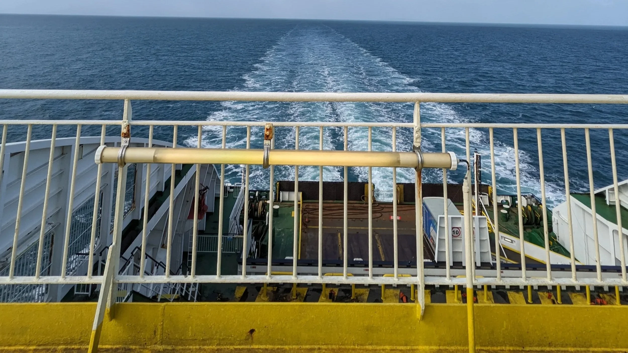 A photo of the sea taken from the back of a boat. There is a trail of white churn behind the ship as it sails. The sky is blue.