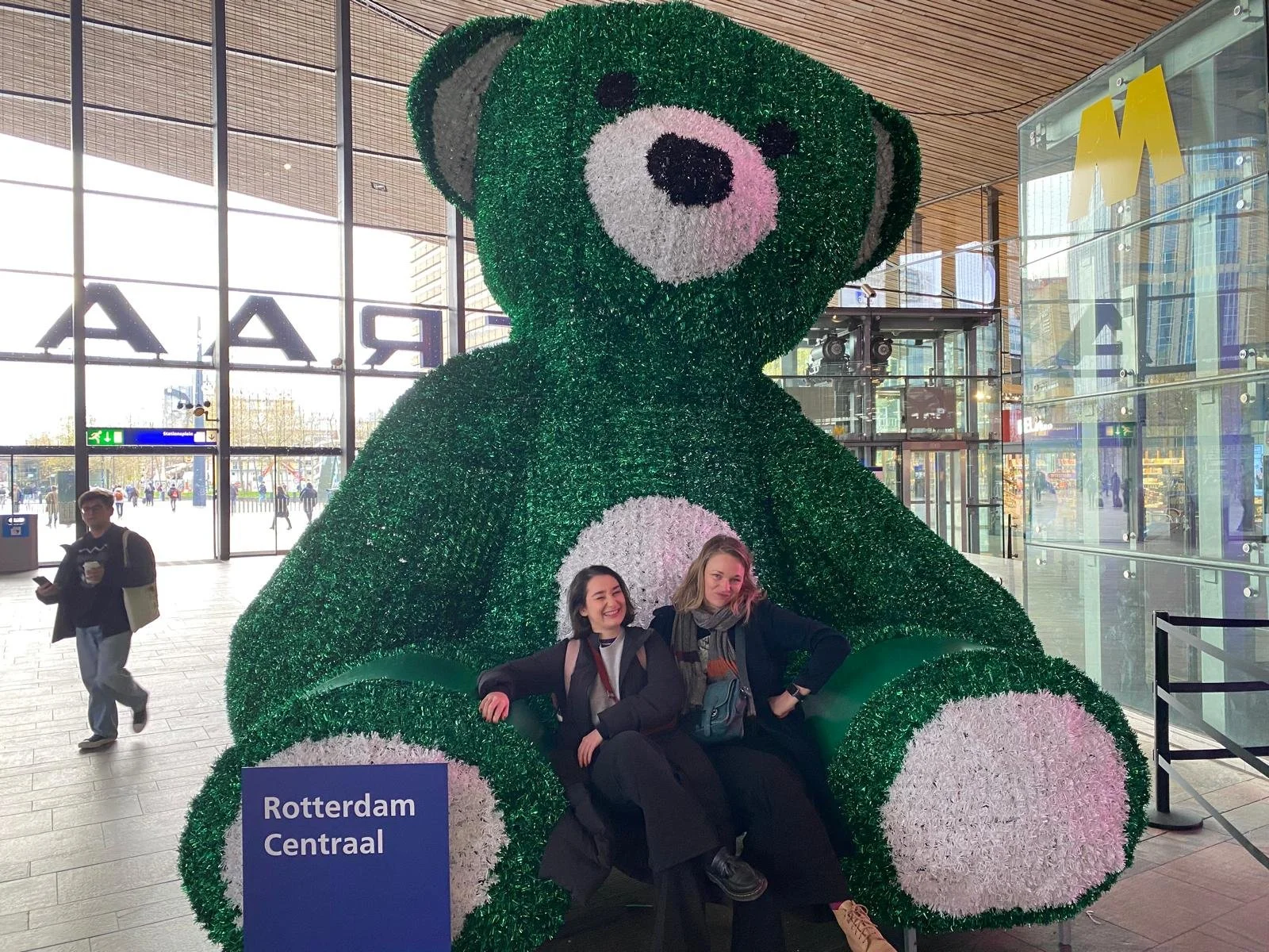 Two women smiling sitting between the legs of a giant green bear statue in Rotterdam train station.