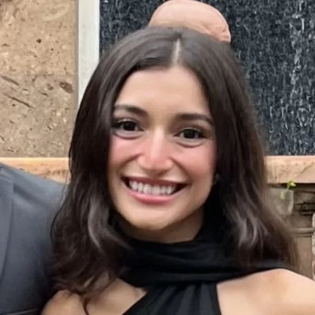 A smiling woman with short brown hair, wearing a gray sweater and a striped scarf, standing indoors next to a wooden wall.