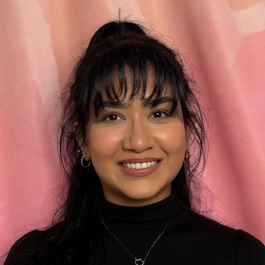 A young woman with dark hair styled in a high ponytail and loose bangs, wearing a black turtleneck, hoop earrings, and a silver necklace, smiling in front of a pink background.