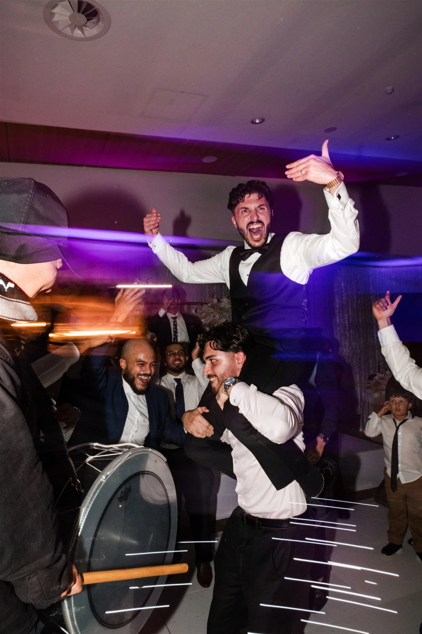 A group of people celebrating at a party, with one man being lifted up and cheering, others dancing and smiling, in a lively indoor setting.