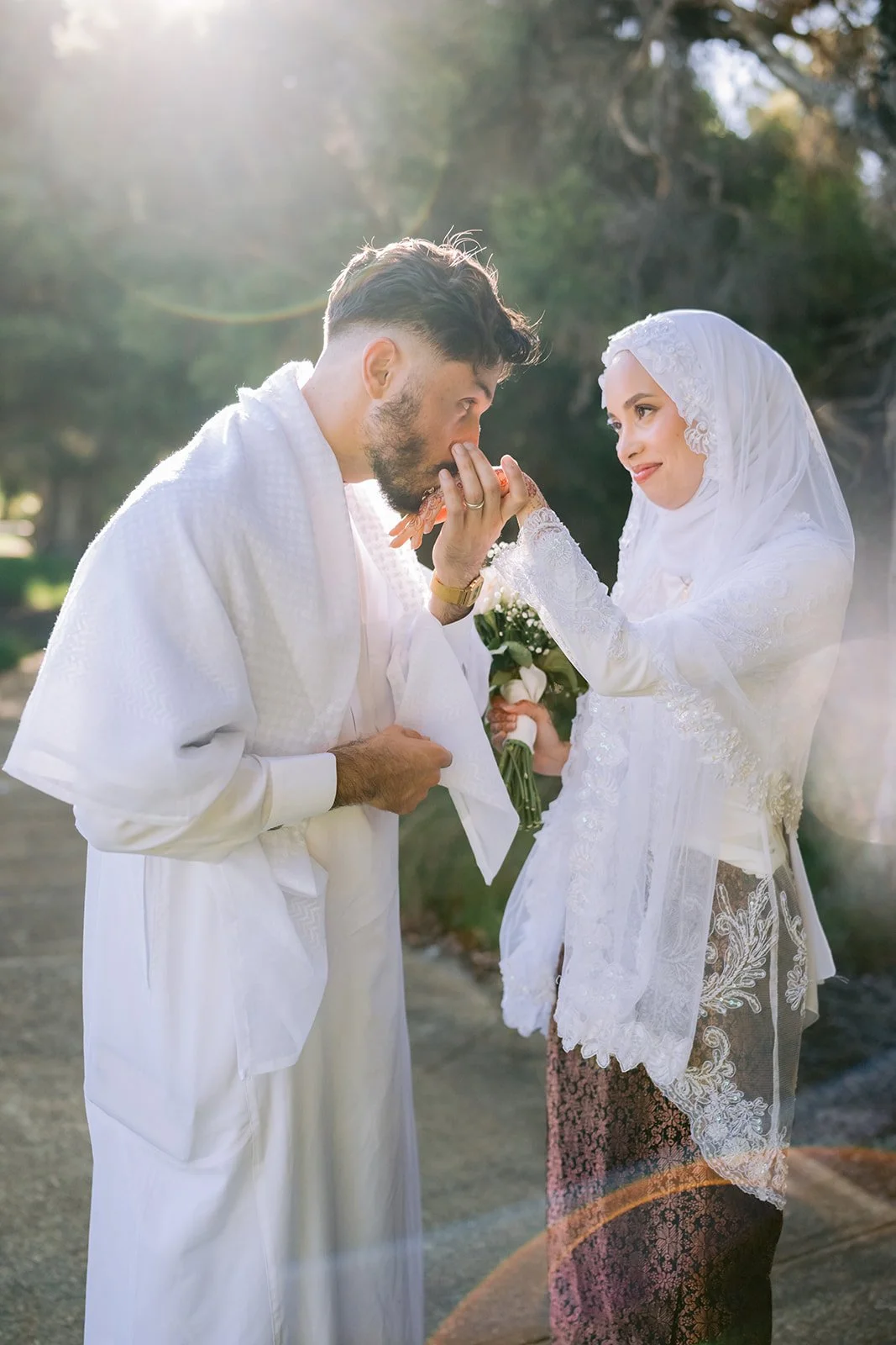 A bride and groom outdoors, with the groom kissing the bride's hand as she holds a bouquet, both dressed in white ceremonial clothing.
