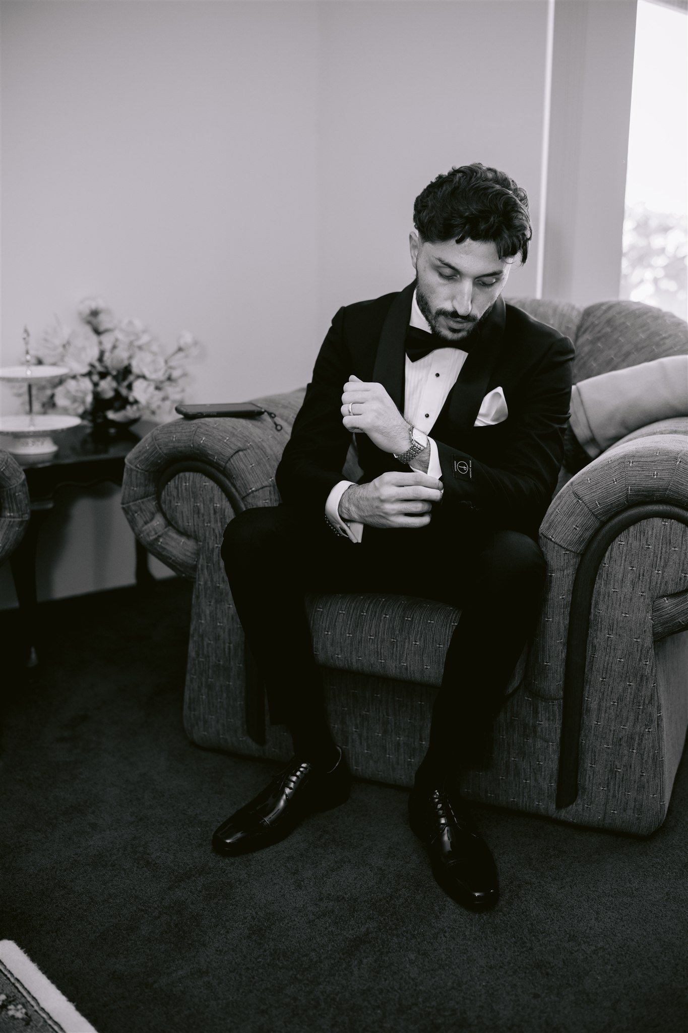 A man in a tuxedo sitting on a sofa, adjusting his cufflinks, in a living room setting with a window and floral arrangement in the background.