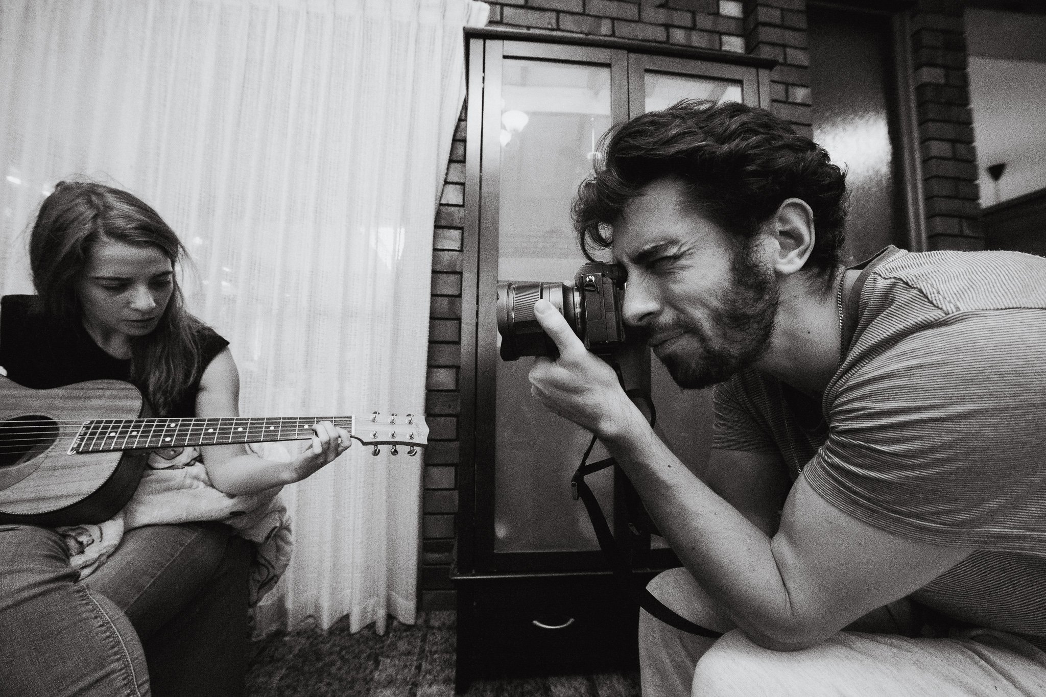 A man taking a photo of a young girl playing guitar indoors in black and white.