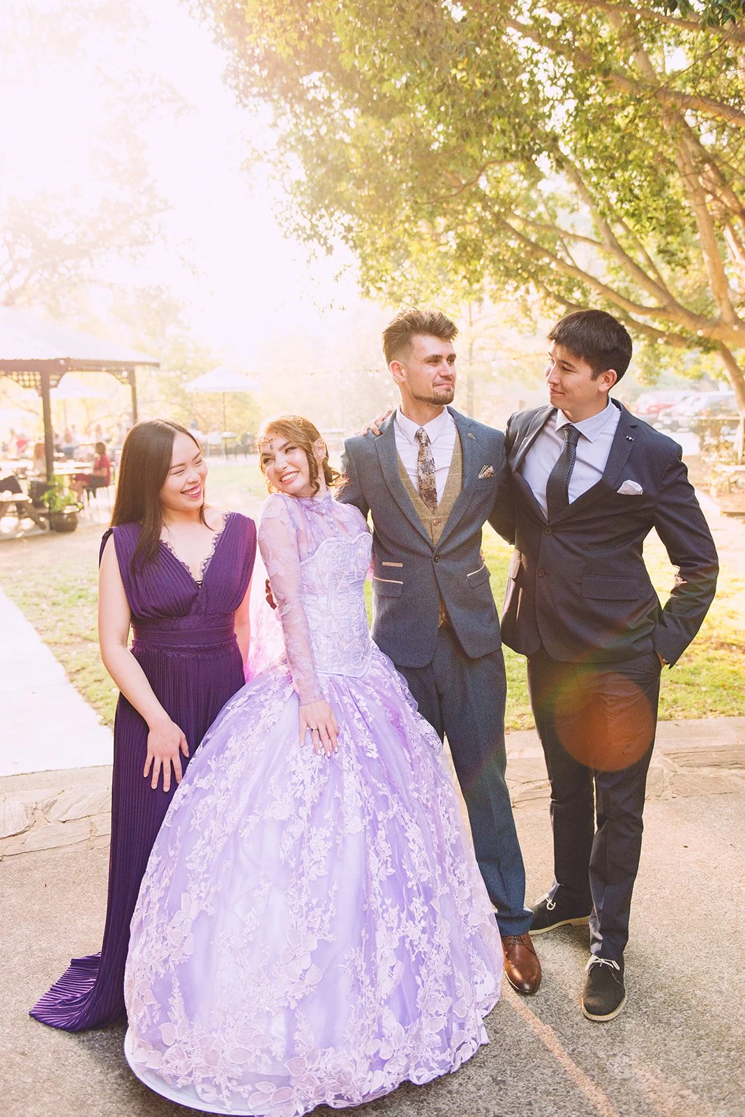 Four young people dressed in formal attire standing outdoors under a tree, smiling and looking at each other, with bright sunlight filtering through the leaves.