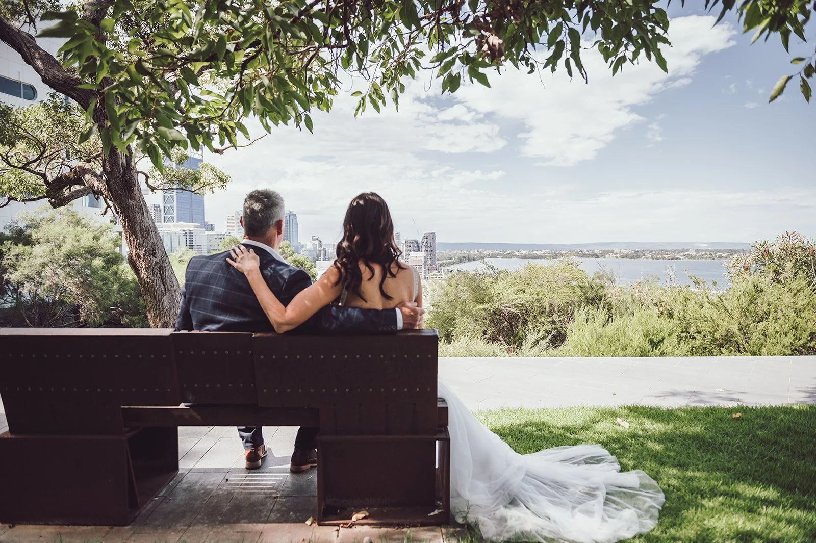 A couple sitting on a bench under a tree, overlooking a city skyline and a river, with the woman wearing a white wedding dress and the man in a suit.