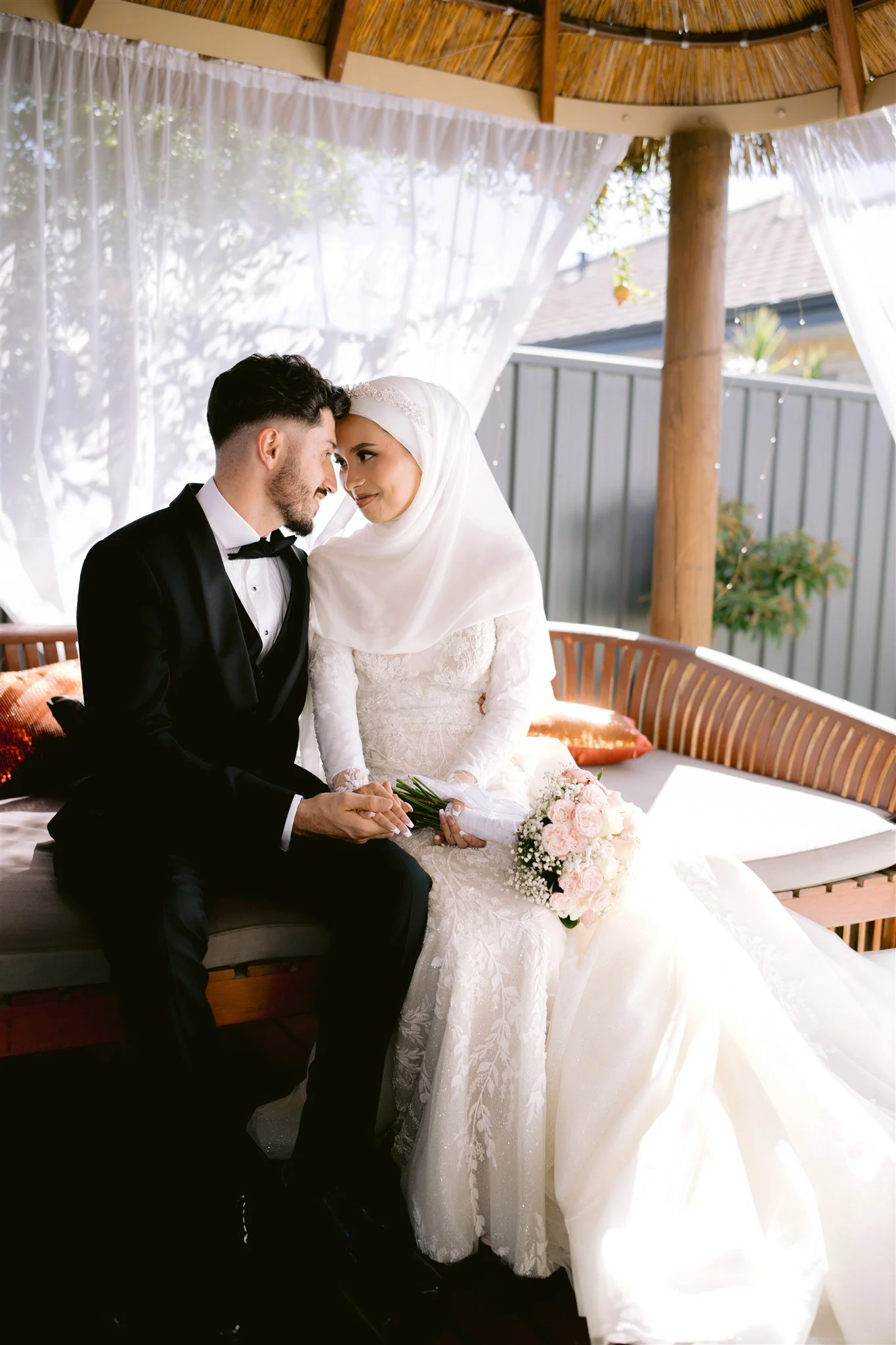 A bride and groom sit closely together on a cushioned outdoor bench, touching foreheads and smiling, with the bride holding a bouquet of pink and white flowers. The groom is dressed in a black tuxedo, and the bride wears a white dress with lace detai