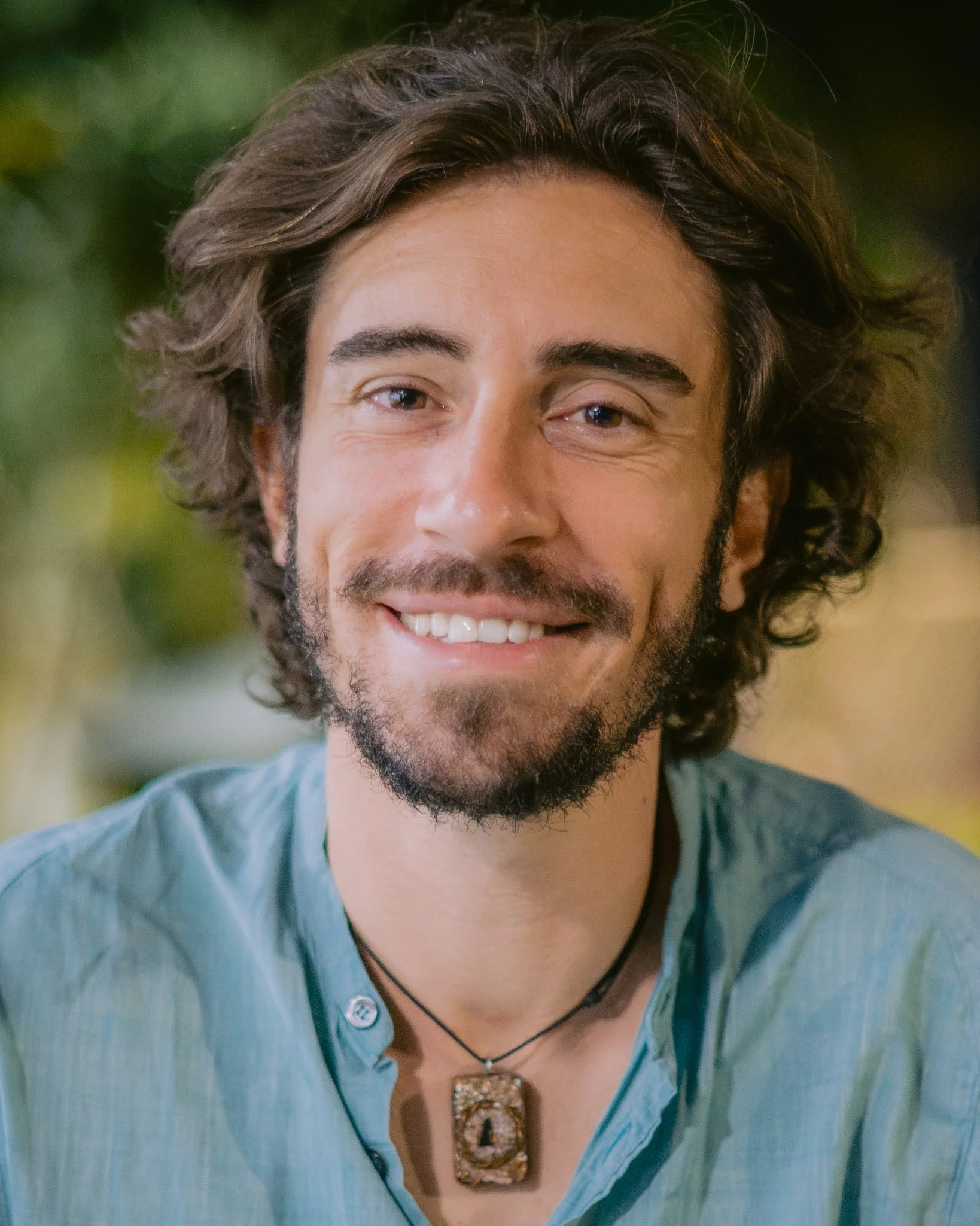 A young man with wavy brown hair, a beard, and a light blue shirt, smiling at the camera, wearing a pendant necklace.
