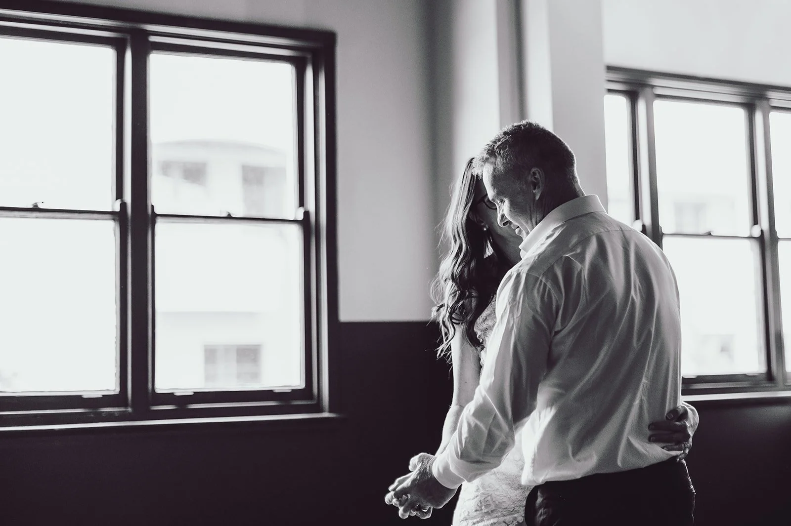 Black and white photo of a man and woman dancing closely indoors near windows, with the man smiling and the woman in glasses, both dressed in formal clothing.