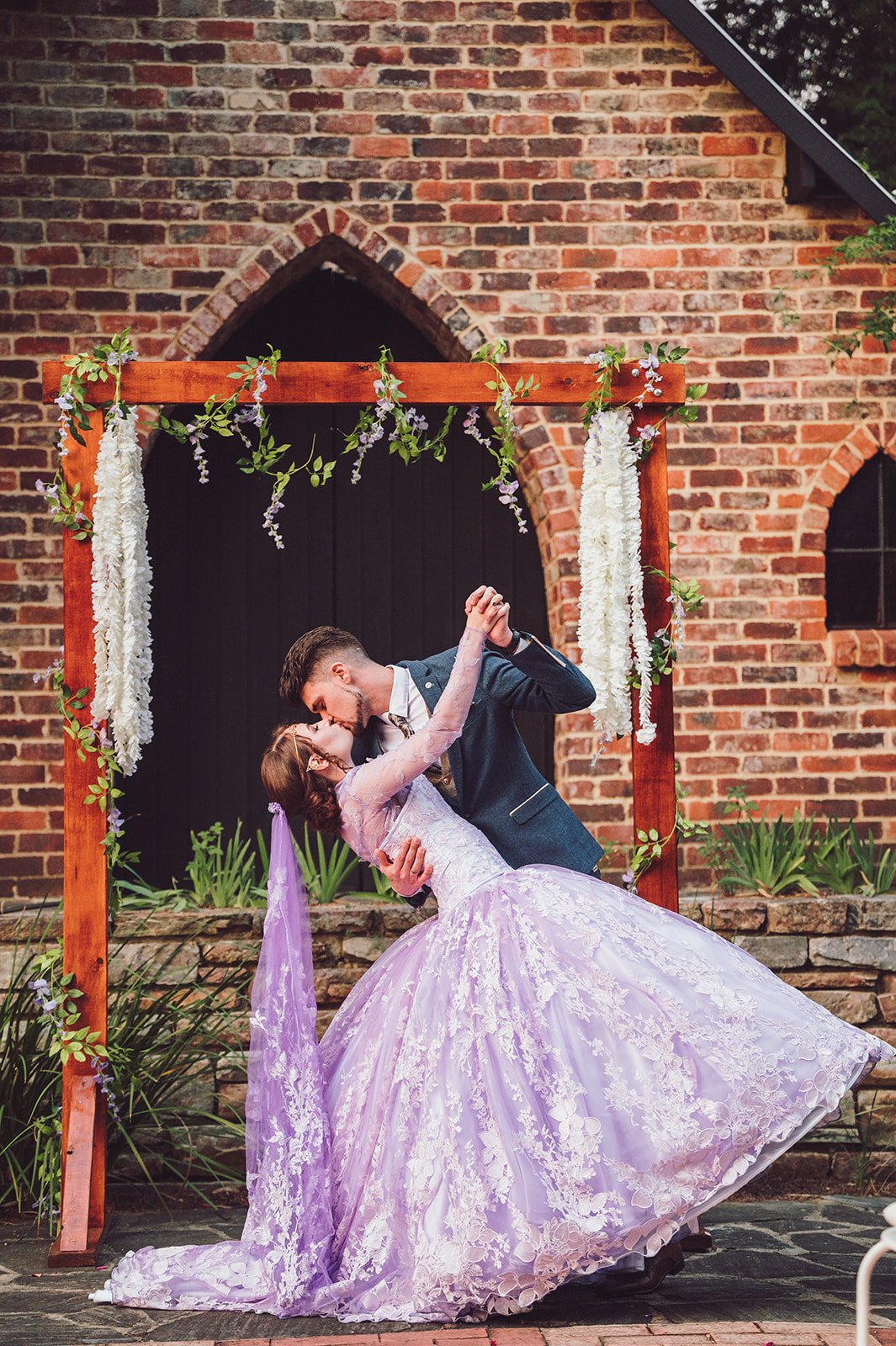 A couple sharing a dance outdoors under a wooden arch decorated with white flowers and greenery, with a brick building in the background.