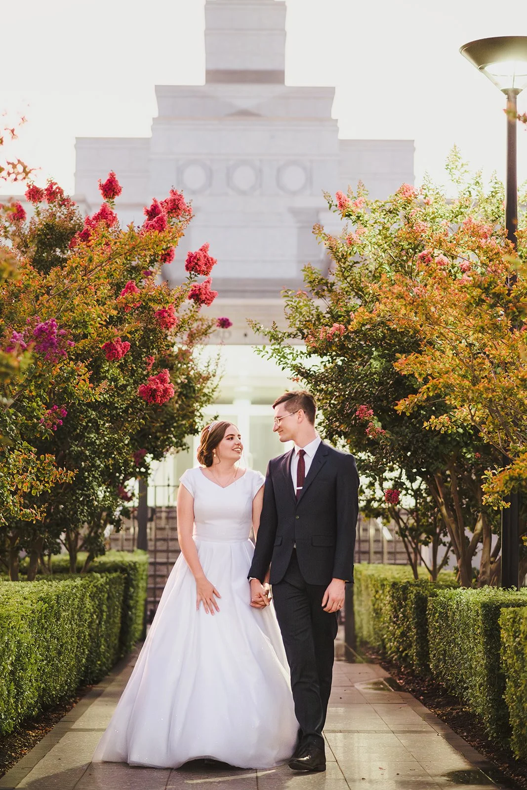 A bride and groom holding hands and walking through a garden with pink and purple flowering trees, with a monument in the background.