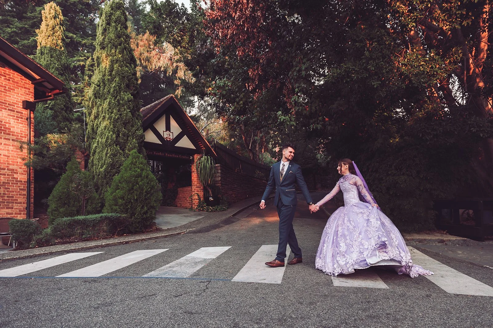 A couple in wedding attire holding hands and crossing a street with a striped crosswalk, surrounded by trees and brick buildings.