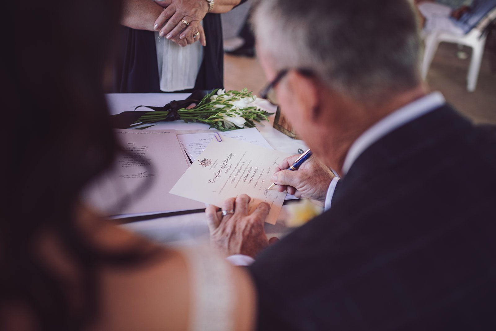 Man in a suit signing a marriage certificate at a wedding ceremony with a bouquet of white flowers on the table.