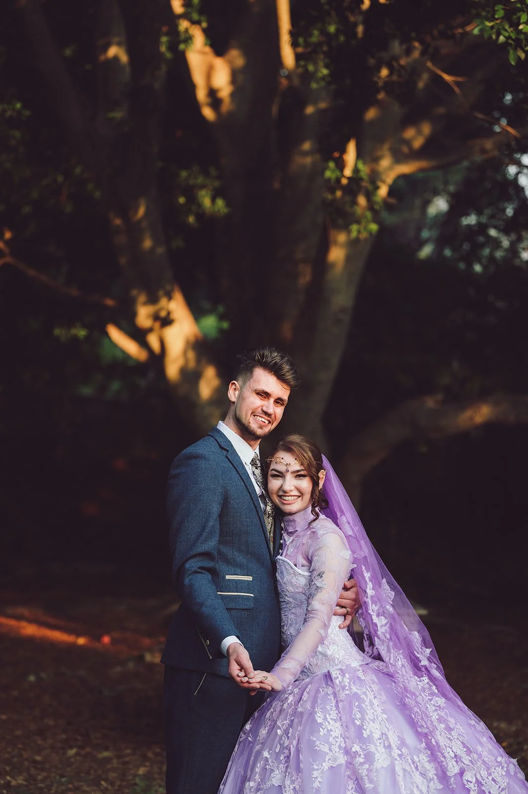 A happy couple dressed in wedding attire, standing outdoors in front of a large tree, smiling and holding hands.