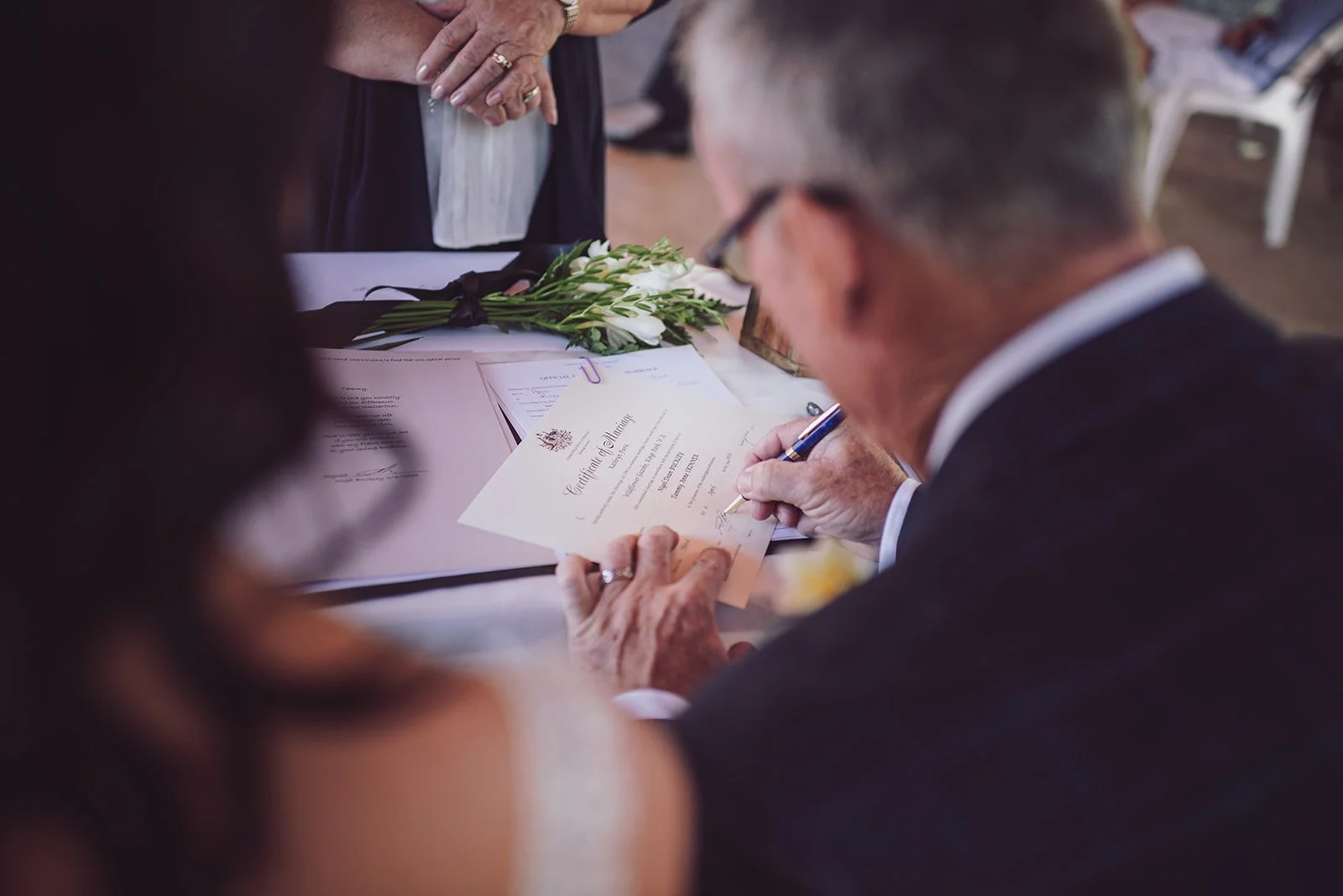 An elderly man in a dark suit signing a marriage certificate at a wedding ceremony.