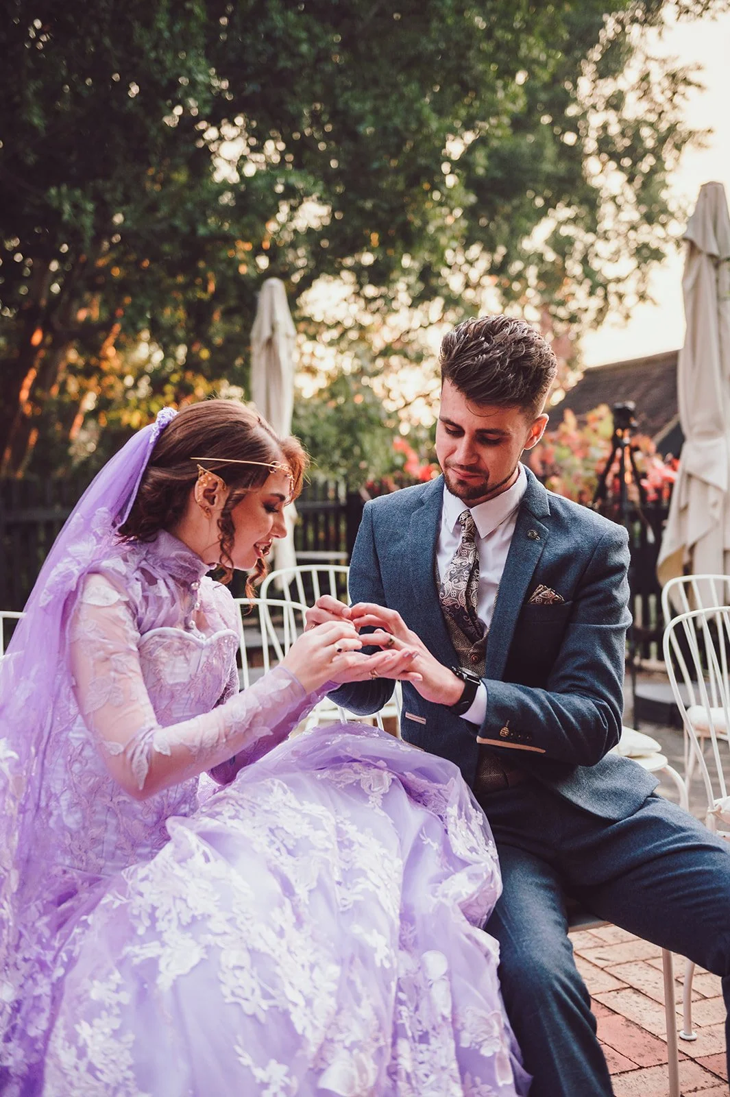 A woman in a lavender gown and veil has a ring placed on her finger by a man in a dark suit, outdoors during sunset with trees and white chairs around.