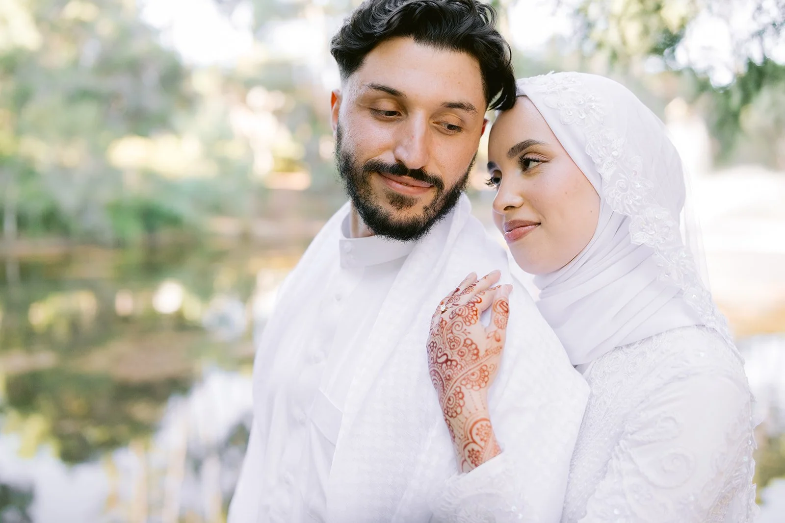 A bride and groom with their foreheads touching in a romantic moment outdoors, the bride wearing a white hijab and dress, the groom in a white shirt, with a background of trees and a pond.