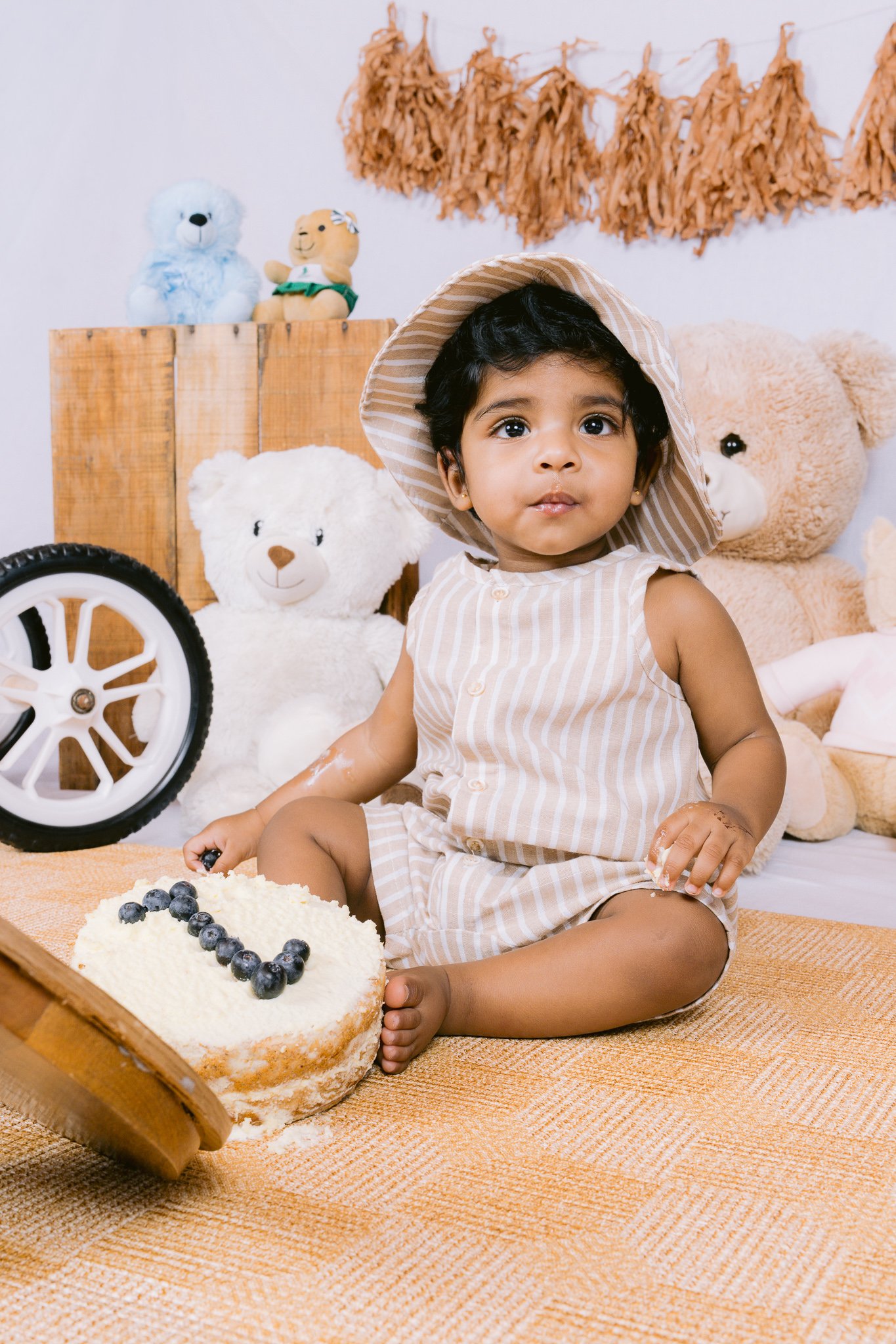 A young girl with dark hair wearing a striped sleeveless dress and a sun hat, sitting on a woven mat with a partially eaten cake decorated with blueberries. She is surrounded by stuffed animals including a large white bear, a large tan teddy bear, and smaller toys in the background.