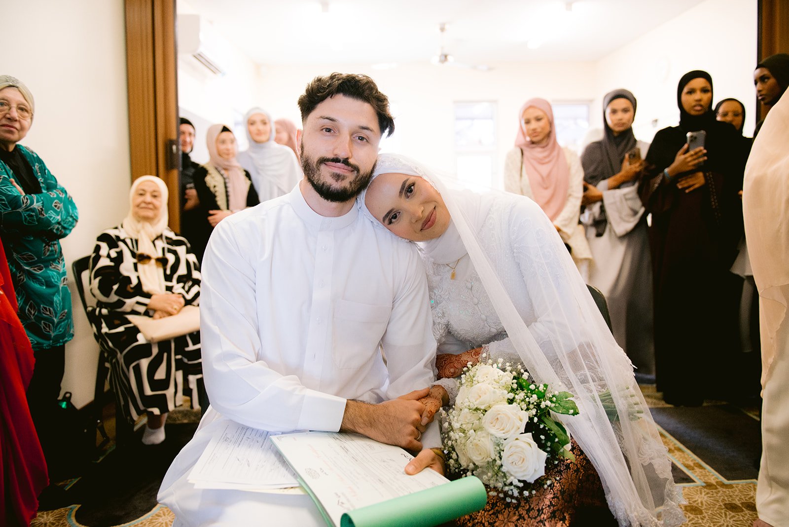 A newlywed couple, a man and a woman in traditional attire, sitting together during their wedding ceremony surrounded by family and friends.