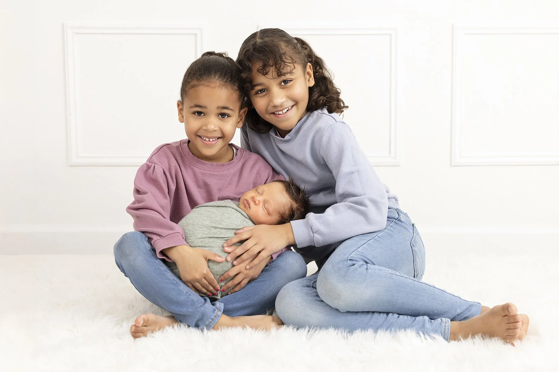 Two kids and a newborn baby sit in a studio on a furry rug