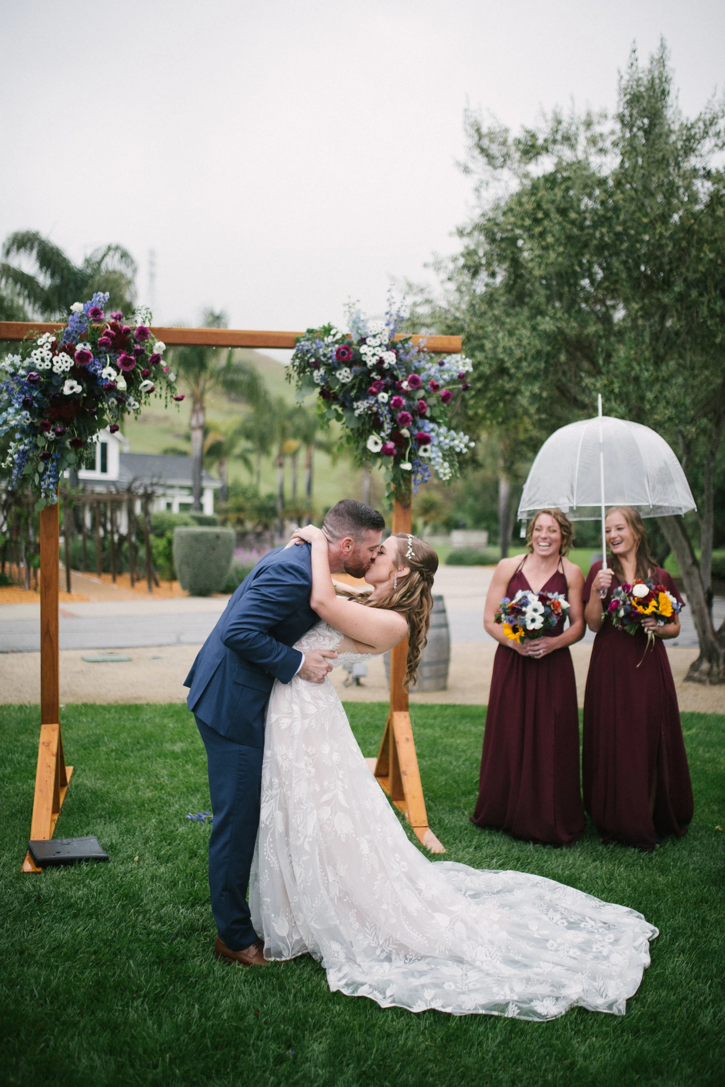 Couple sharing a kiss during a wedding ceremony outdoors under a floral arch, with two bridesmaids holding bouquets and an umbrella standing nearby.