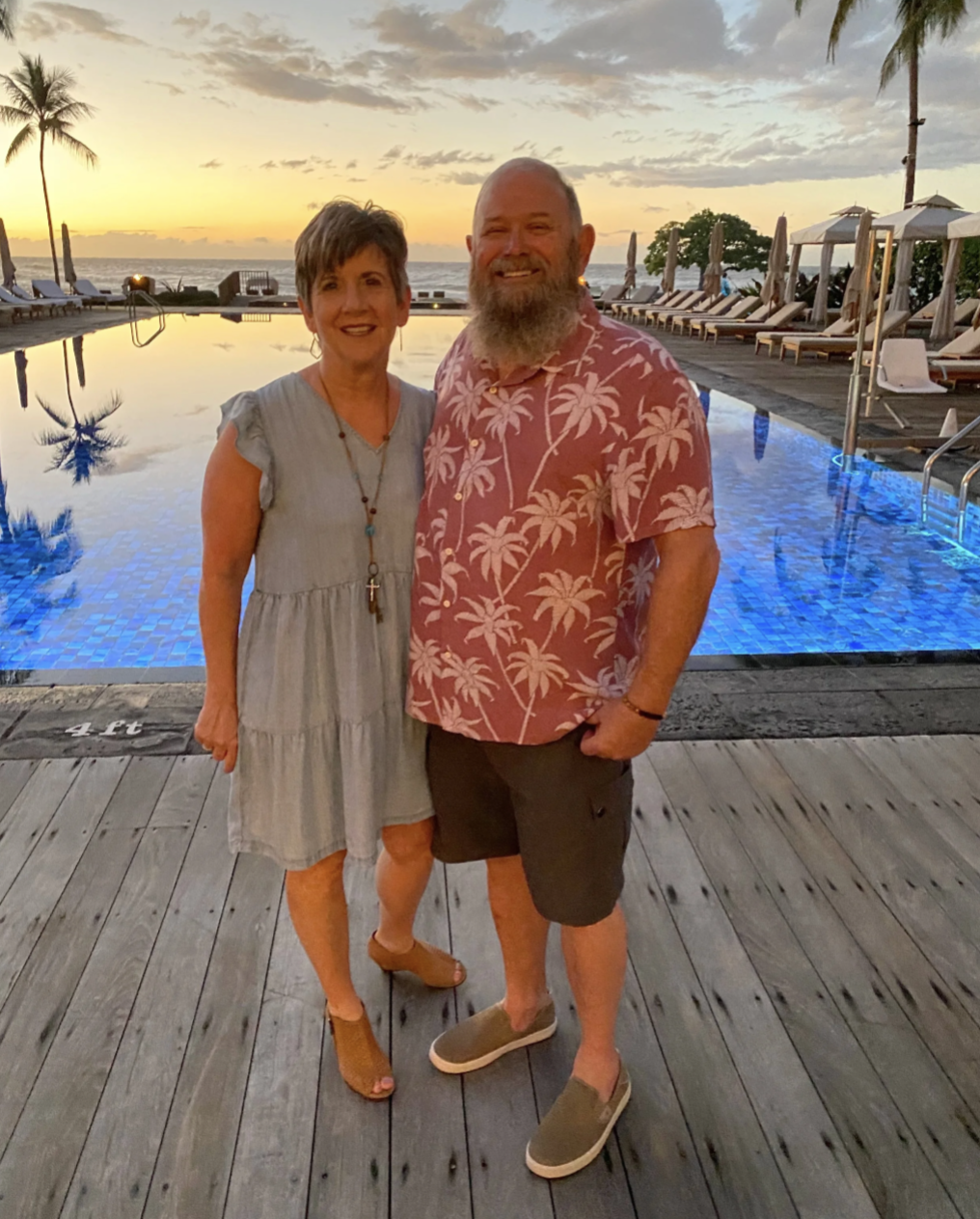 A smiling couple stands by a pool at sunset, with a tropical ocean backdrop and palm trees, surrounded by lounge chairs and umbrellas.