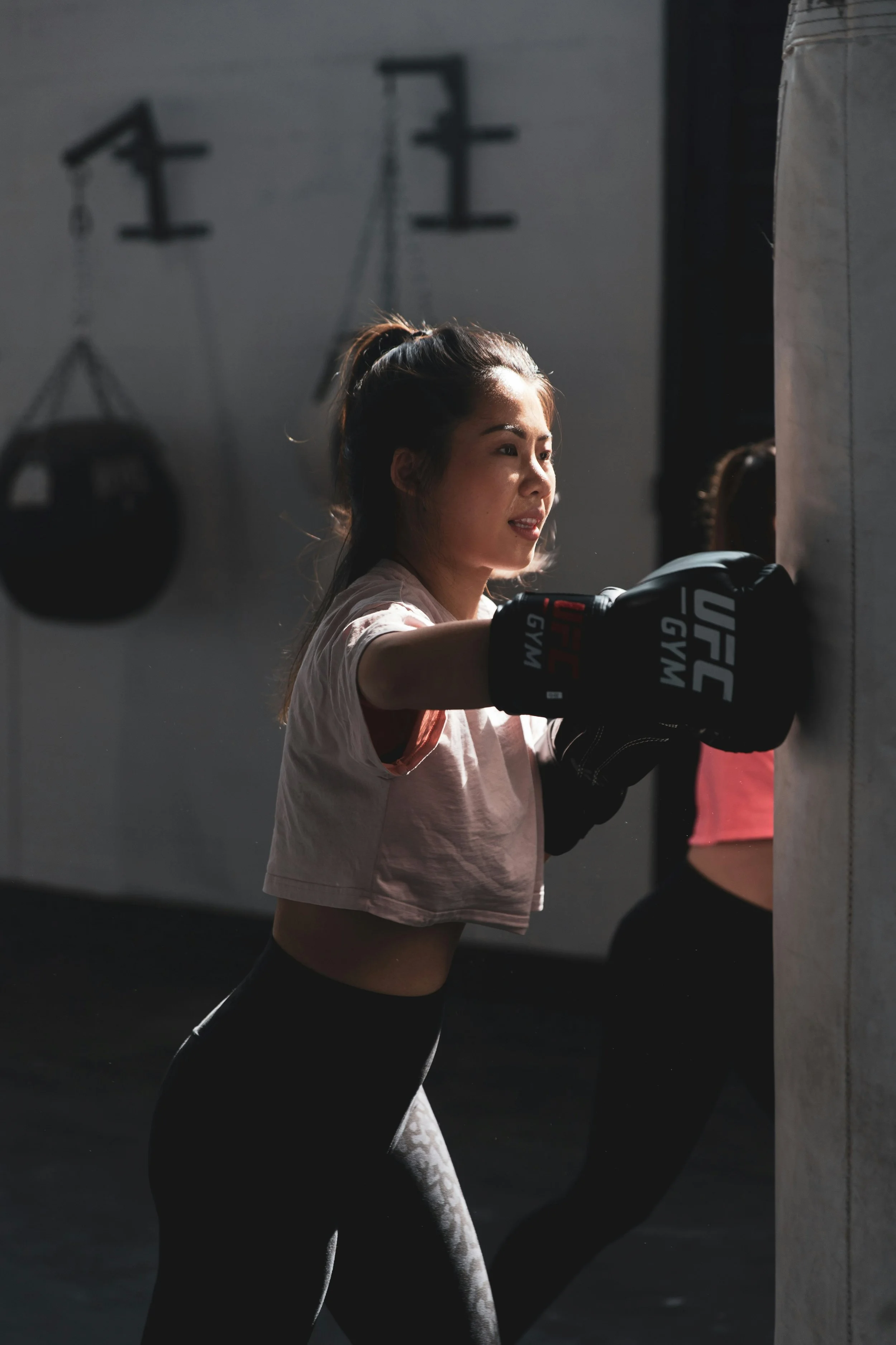 A woman with a ponytail and sports gloves is punching a boxing bag in a gym, with weighing equipment and a pole in the background.