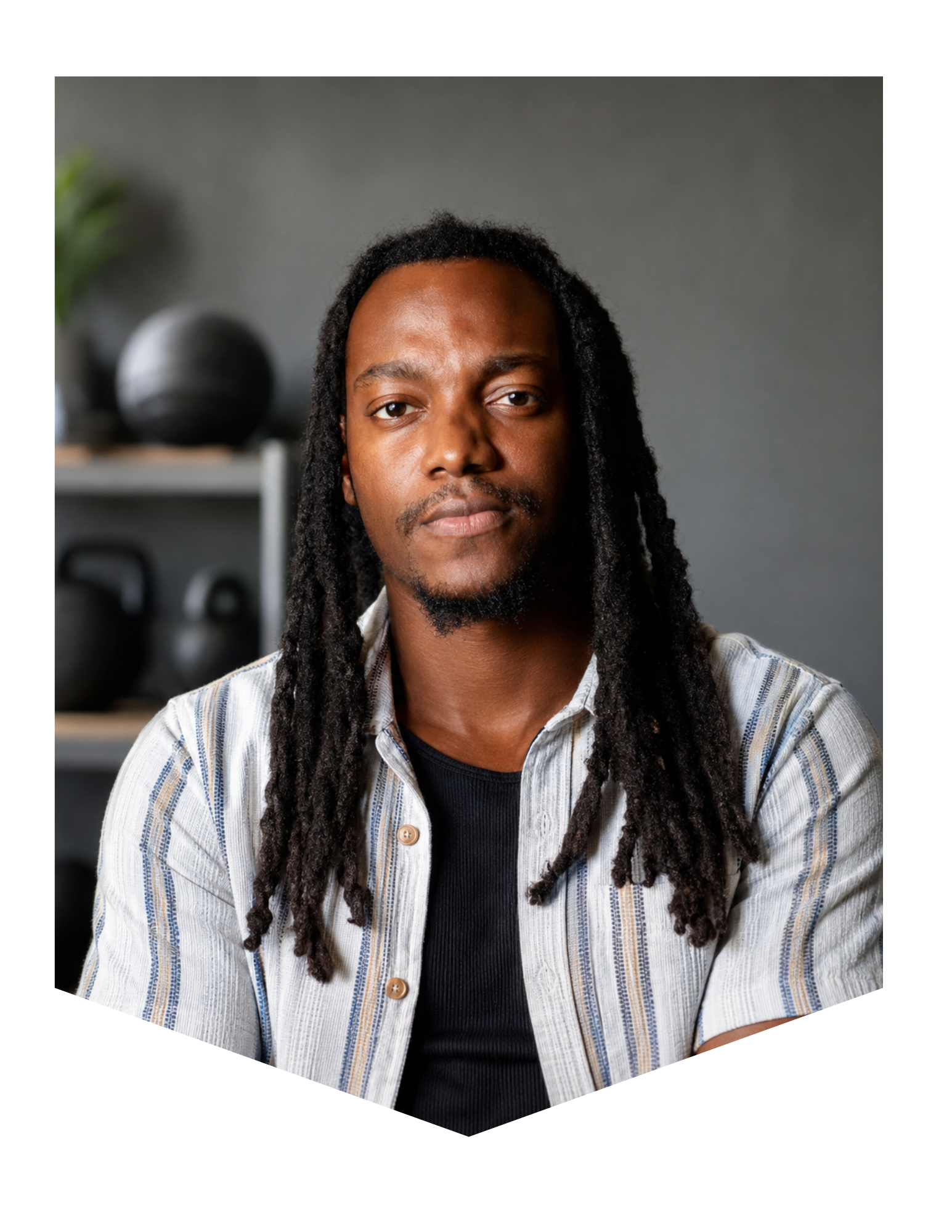 Portrait of a young man with long dreadlocks, wearing a striped button-up shirt over a black t-shirt, sitting in front of a gray background with shelving and decorative objects.