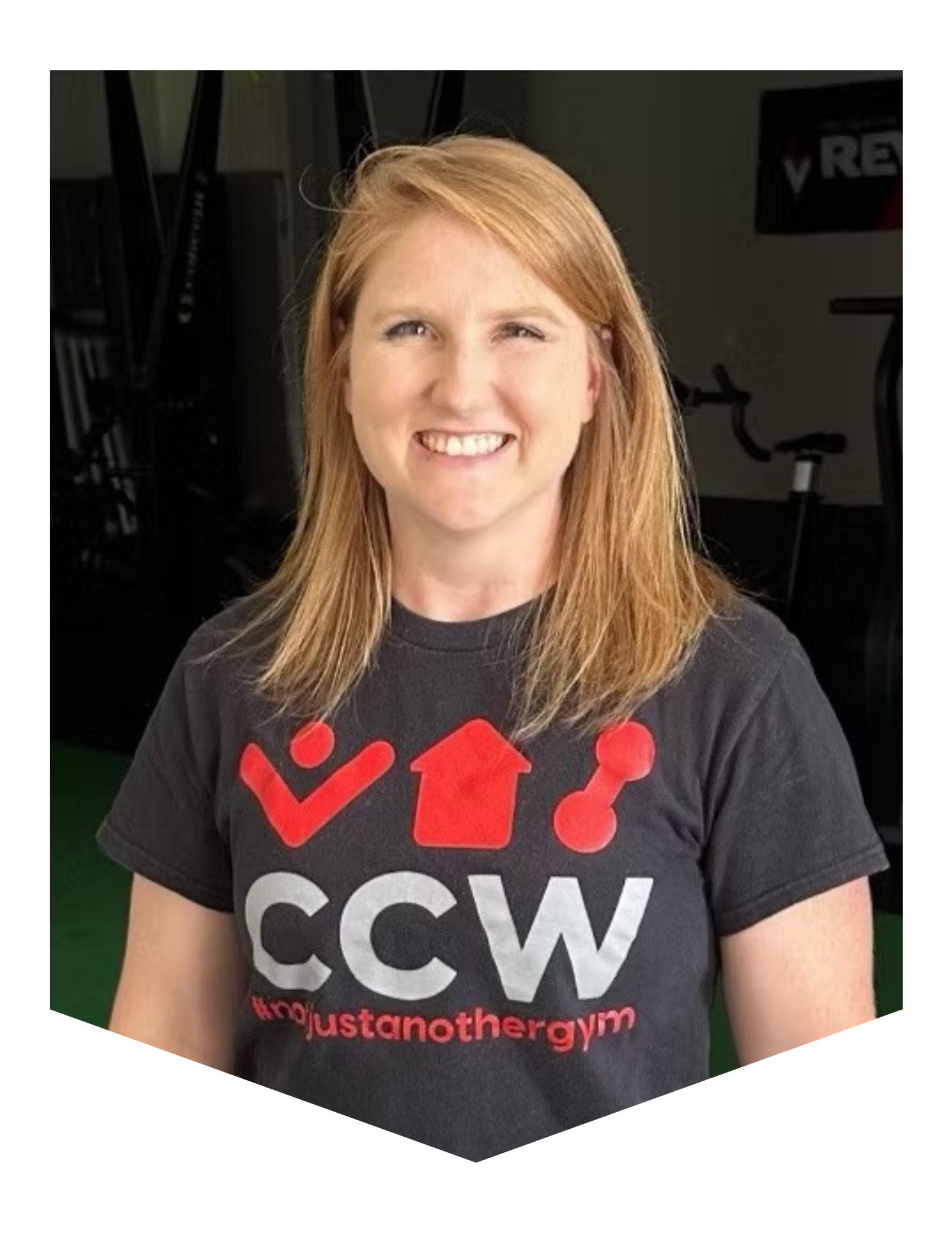 A smiling woman with red hair wearing a black T-shirt with red and white text and symbols supporting a cause, in a gym setting.