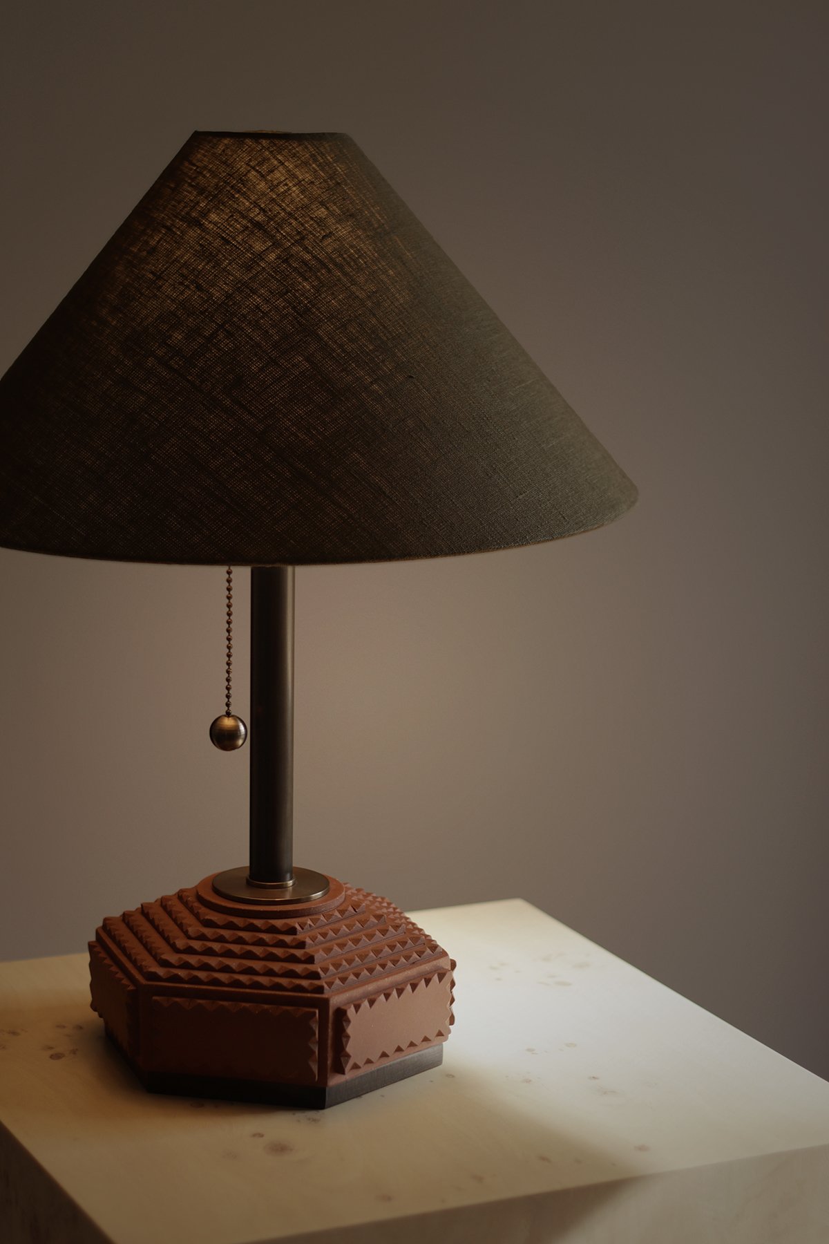 An artist made table lamp with a dark, conical shade and a textured, hexagonal base sits on a light wooden surface. Soft lighting creates a warm, cozy ambiance.