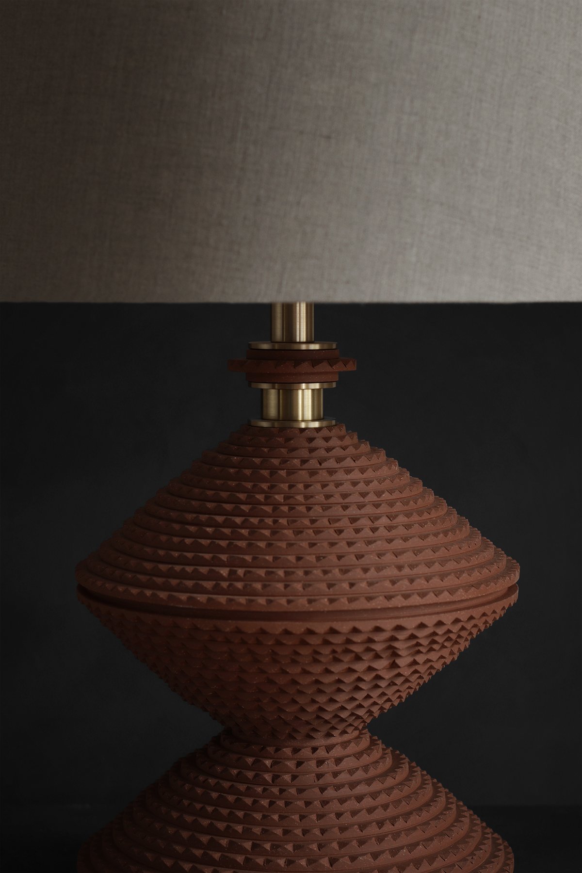 Close-up of a modern table lamp with a textured, angural, terracotta base and a brown linen fabric shade against a dark background, conveying elegance.