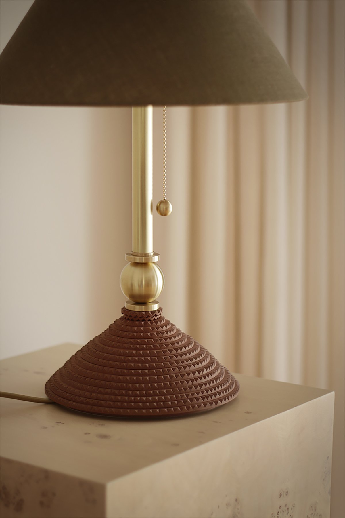 A hand made table lamp with a dark green shade, ceramic base and tall brass neck, on a wooden pedestal. Soft light and neutral curtains create a calming, elegant atmosphere.