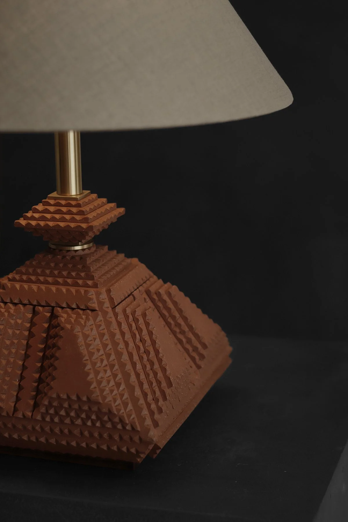 A handcrafted, geometric table lamp with a warm brown base and a beige fabric shade sits on a dark stand against a black background, creating a cozy ambiance.