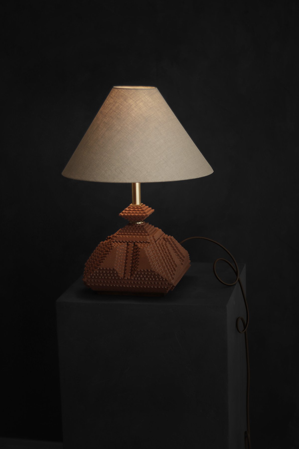 A handcrafted, geometric table lamp with a warm brown base and a beige fabric shade sits on a dark stand against a black background, creating a cozy ambiance.