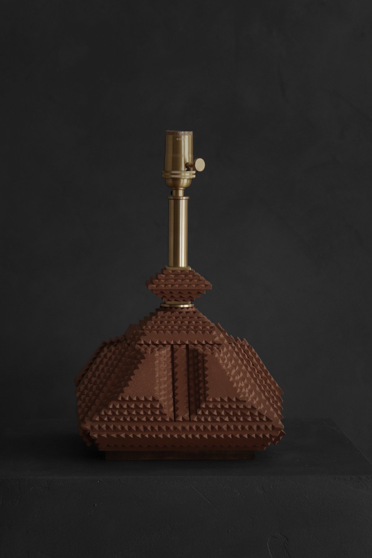 A handcrafted, geometric table lamp with brass details sits on a dark stand against a black background, creating a cozy ambiance.