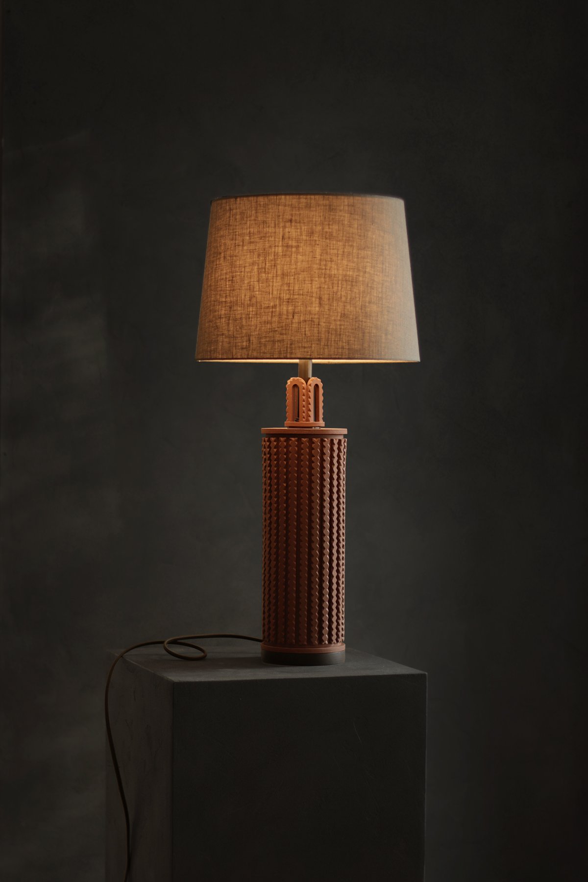 Illuminated hand carved terracotta clay table lamp with a brown shade on a gray pedestal against a dark background. It emits a warm, soft glow, creating a cozy ambiance.