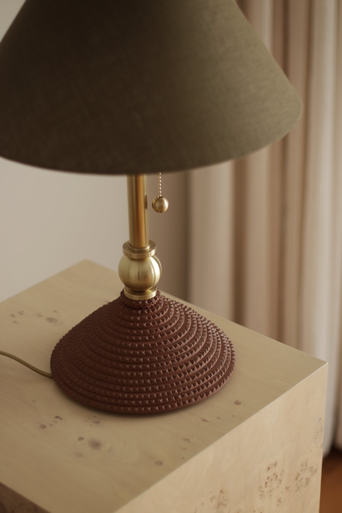 A hand made table lamp with a dark green shade, ceramic base and tall brass neck, on a wooden pedestal. Soft light and neutral curtains create a calming, elegant atmosphere.
