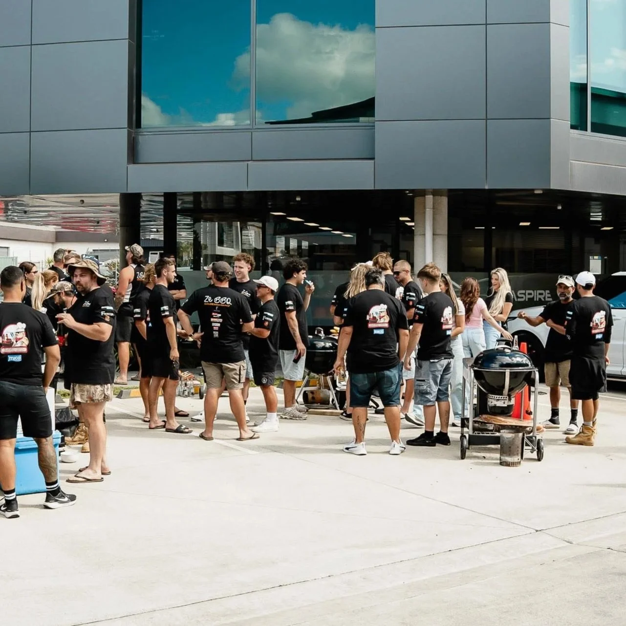 People gathered outside a modern building for a barbecue event, many wearing black t-shirts, some with grilling equipment, socializing and preparing food.