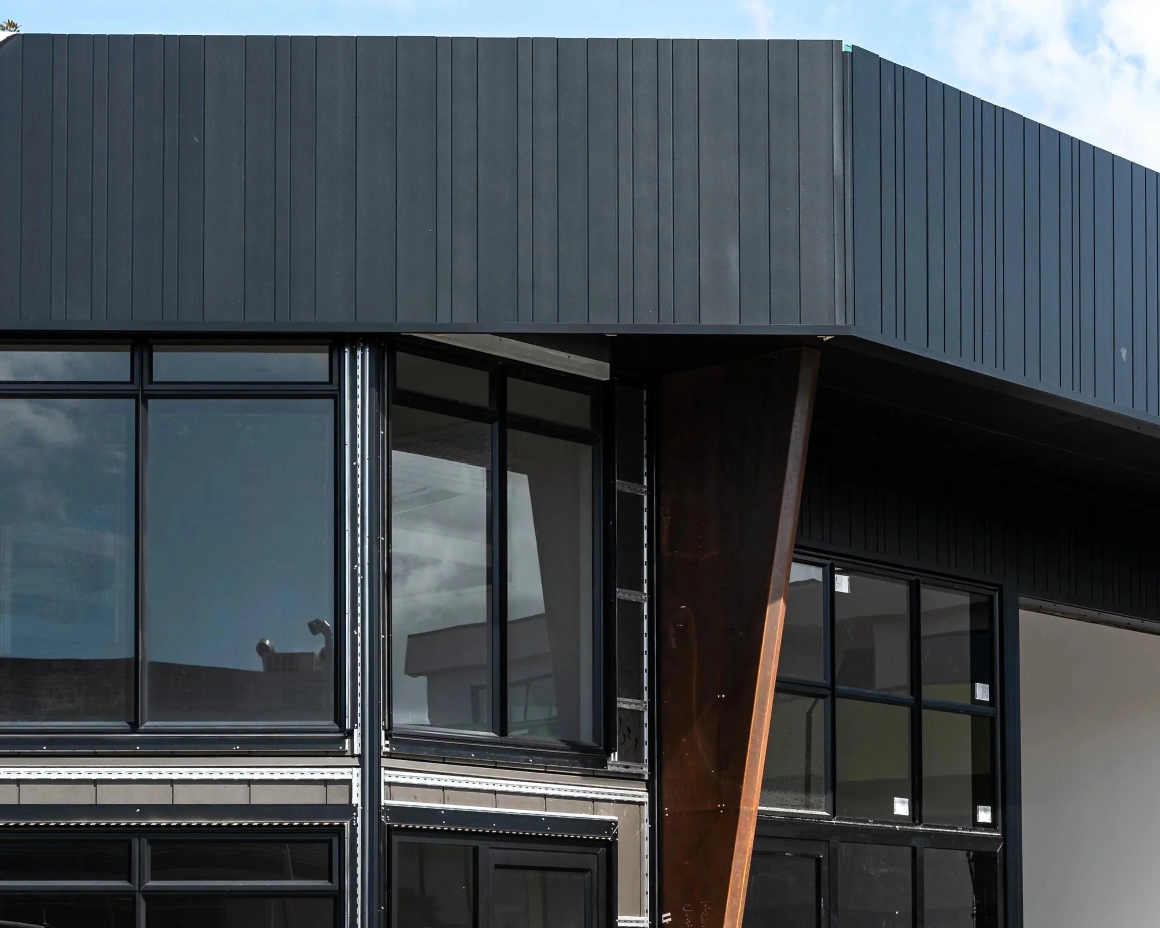 Close-up of a modern architectural building featuring dark metal paneling and large glass windows, with a cloudy sky in the background.