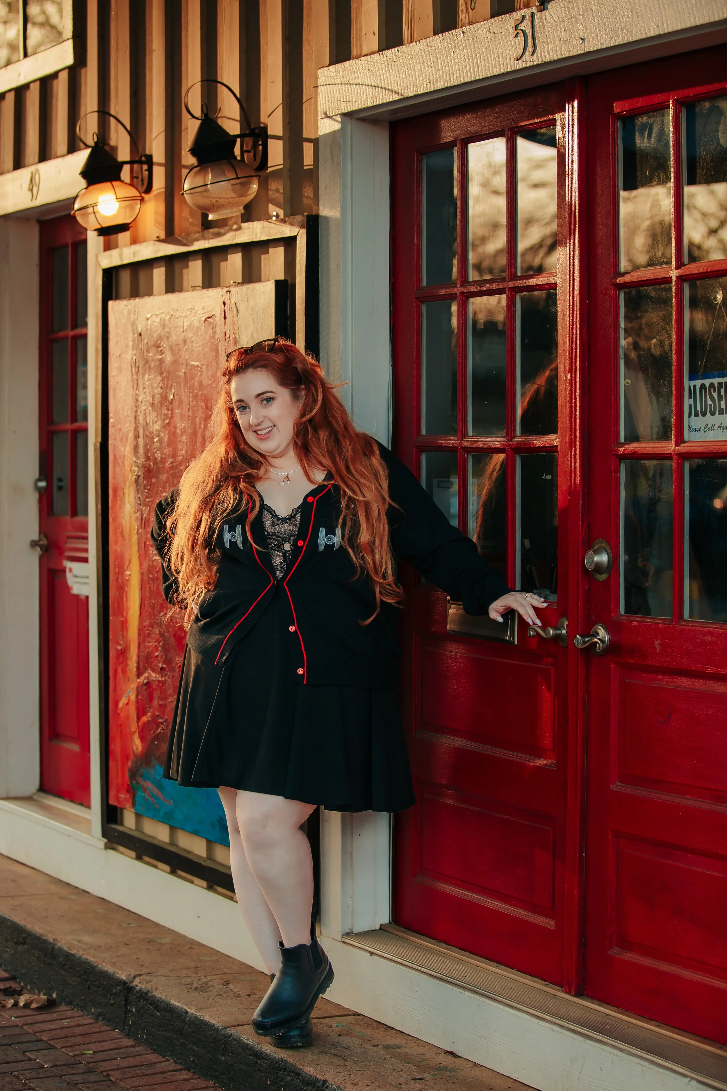 A young woman with long red hair standing outside a red door of a building, smiling at the camera, wearing black clothing with red accents and black boots.