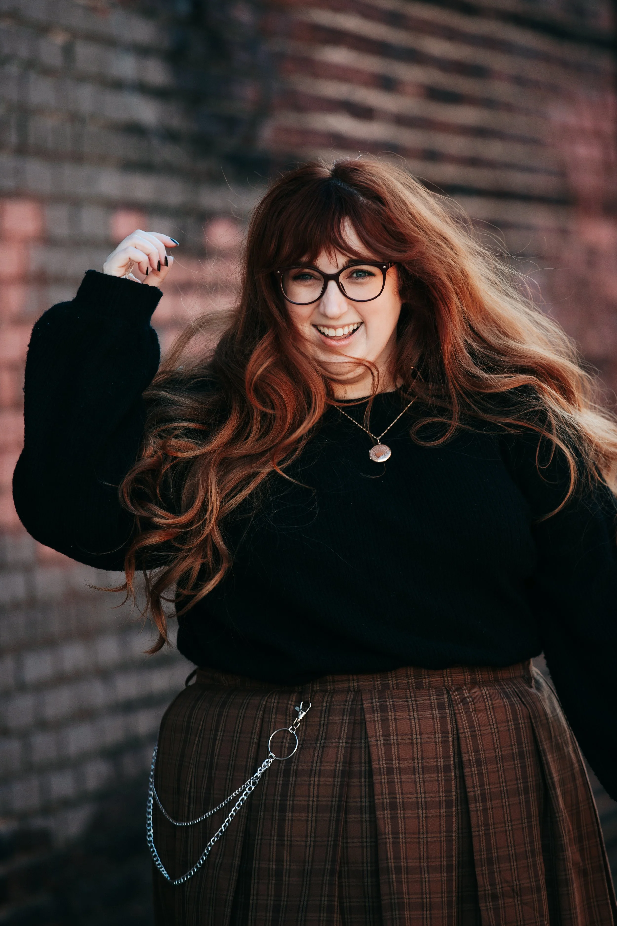 A woman with long, wavy red hair and glasses, smiling and waving, standing next to a brick wall.
