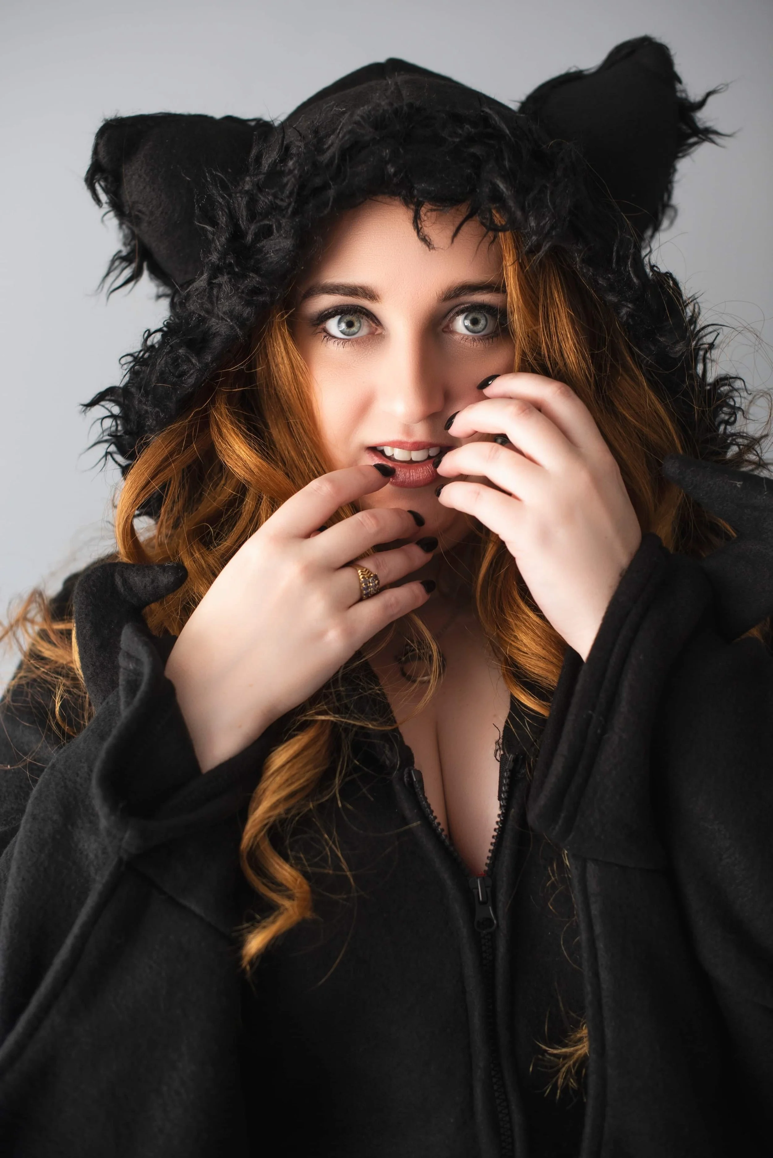 A young woman with long red hair and blue eyes wearing a black hoodie and black gloves, looking at the camera with a playful expression, with her hands near her face.