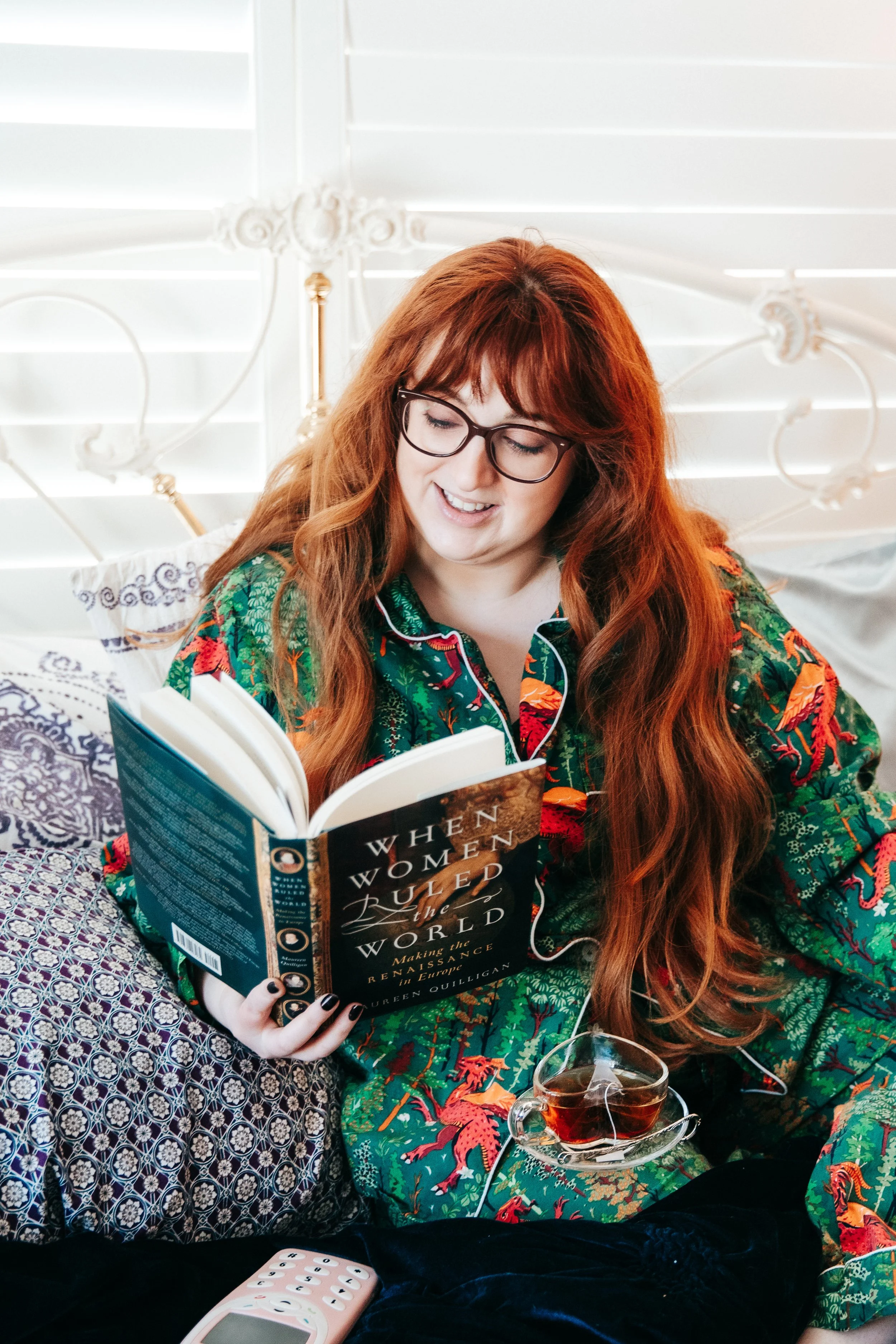 Woman with long red hair and glasses sitting on bed, reading a book titled 'When Women Ruled the World,' with a glass of tea nearby.