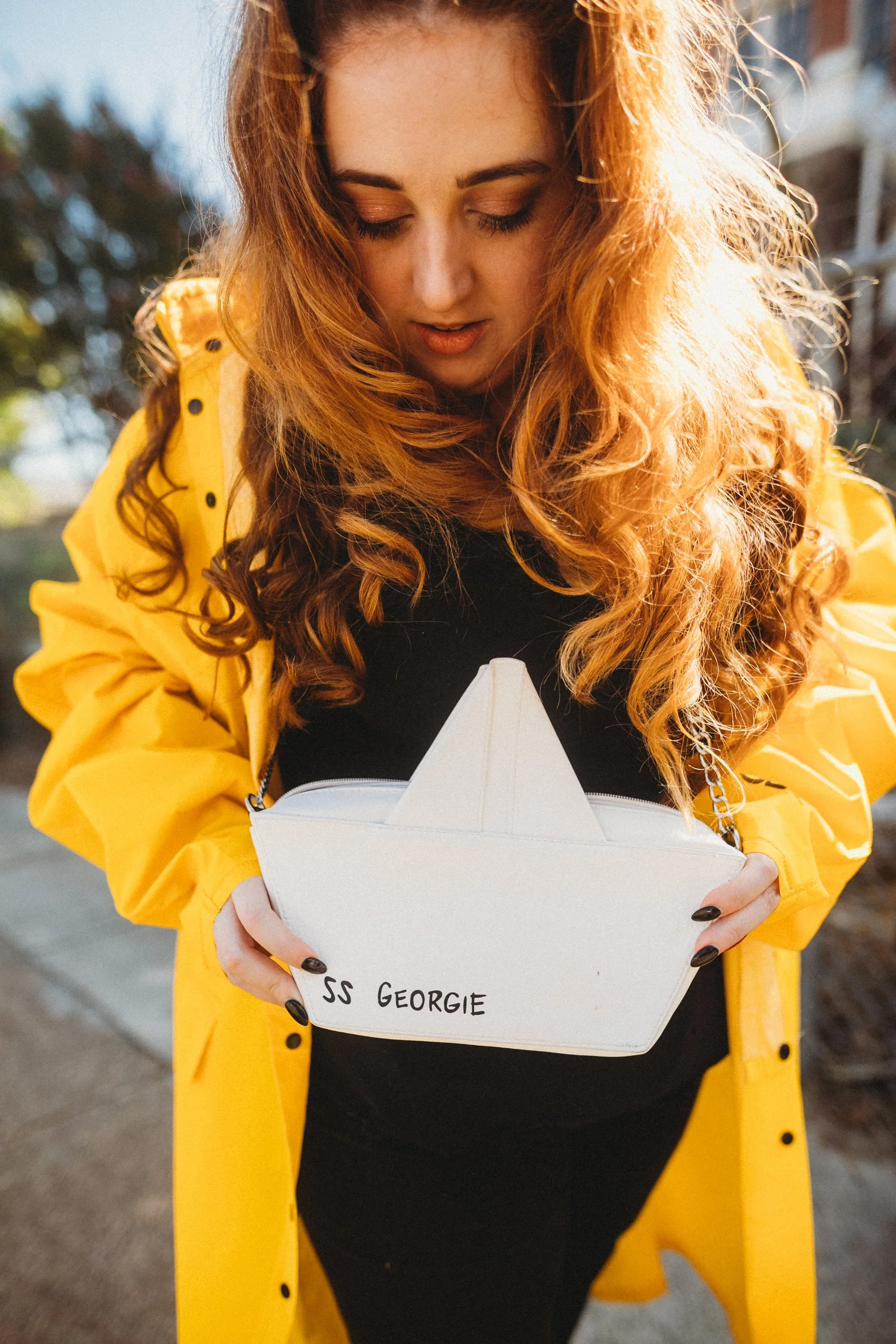 A woman with long, curly red hair wearing a yellow jacket looking into a white purse shaped like a boat with black handwritten text, outdoors with trees in the background.