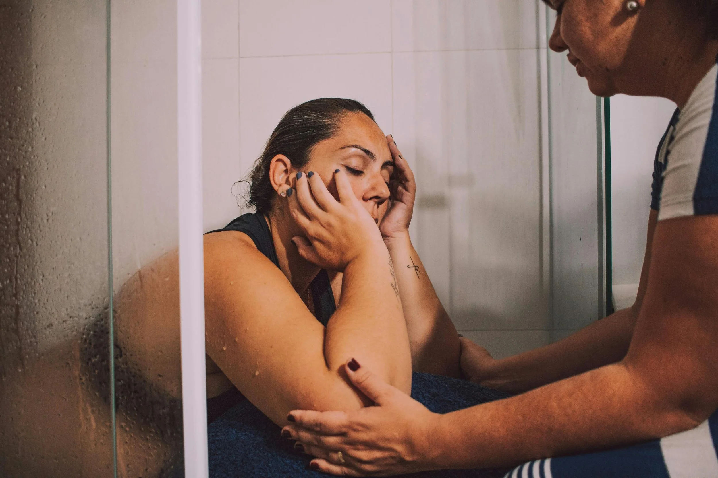 A woman sitting in a shower, appearing distressed, as a caregiver offers comfort and support.